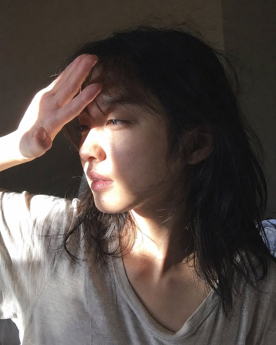 Nine_Shell's tweet photo. The shoulder is a little off in this one, but I’m letting it slide — I’ve already done too many rerolls.

Here’s a shorter version of the prompt. It loses a bit of precision, but it should still get you close.

Prompt:

An unusually beautiful fictional adult 20-year-old Japanese woman in an off-duty everyday moment, captured as a casual handheld rear-smartphone photo, not a selfie. Early morning in a dark apartment bedroom, all lights off, door closed. A single narrow slit of low morning sunlight enters almost horizontally from the left through an unseen gap in blackout curtains outside the frame. The window, curtains, gap, sky, and exterior are not visible. The sunlight is seen only by its effect as one narrow hard-edged patch of light, not as a visible beam in the air.

She has just woken up and is sitting upright near the pillow, wearing a worn oversized T-shirt used as sleepwear, with a slightly stretched collar, messy sleep-flattened hair, and a completely bare face. A separate unseen photographer is seated opposite her at the near edge of the bed, about 80–100 cm away, using the rear camera of a smartphone from a natural face-to-face distance, held horizontally slightly below her eye level. The shot is a close bust-up portrait. Most of the bed, bedding, and room are cropped out or nearly invisible.

Her face turns toward the left-side sunlight, creating a left-facing three-quarter view close to profile. She keeps her left hand raised near her brow to soften the incoming light. Only the left hand, wrist, and a small part of the forearm are visible. The hand partly breaks the slit-shaped sunlight before it reaches her face, so the light falls as an irregular narrow hard-edged patch across the side of her hand, fingers, brow, cheekbone, nose edge, lips, and part of the lit side of her face.

She looks freshly awake, sleepy, and mildly disoriented, not posed. She does not look upward. Her head stays level, her chin remains neutral or slightly lowered, and her gaze is horizontal, unfocused, and not directed at the lens. The phone exposes for the harsh sunlit skin, so the brightest lit areas may clip slightly, while the shadow side falls into deep dark tones with subtle digital noise. Her visible skin should feel physically real and unpolished: visible pores, slight uneven skin tone, faint under-eye darkness, subtle redness, natural dry lips, mildly imperfect lived-in hair, tiny imperfections, and tactile morning texture. She should remain obviously beautiful, but without glamour, heavy makeup, whitening, perfect composition, cinematic lighting, polished hair, doll-like features, professional posing, pin-up posing, fetish framing, hidden-camera 느낌, anime style, text, or watermark.