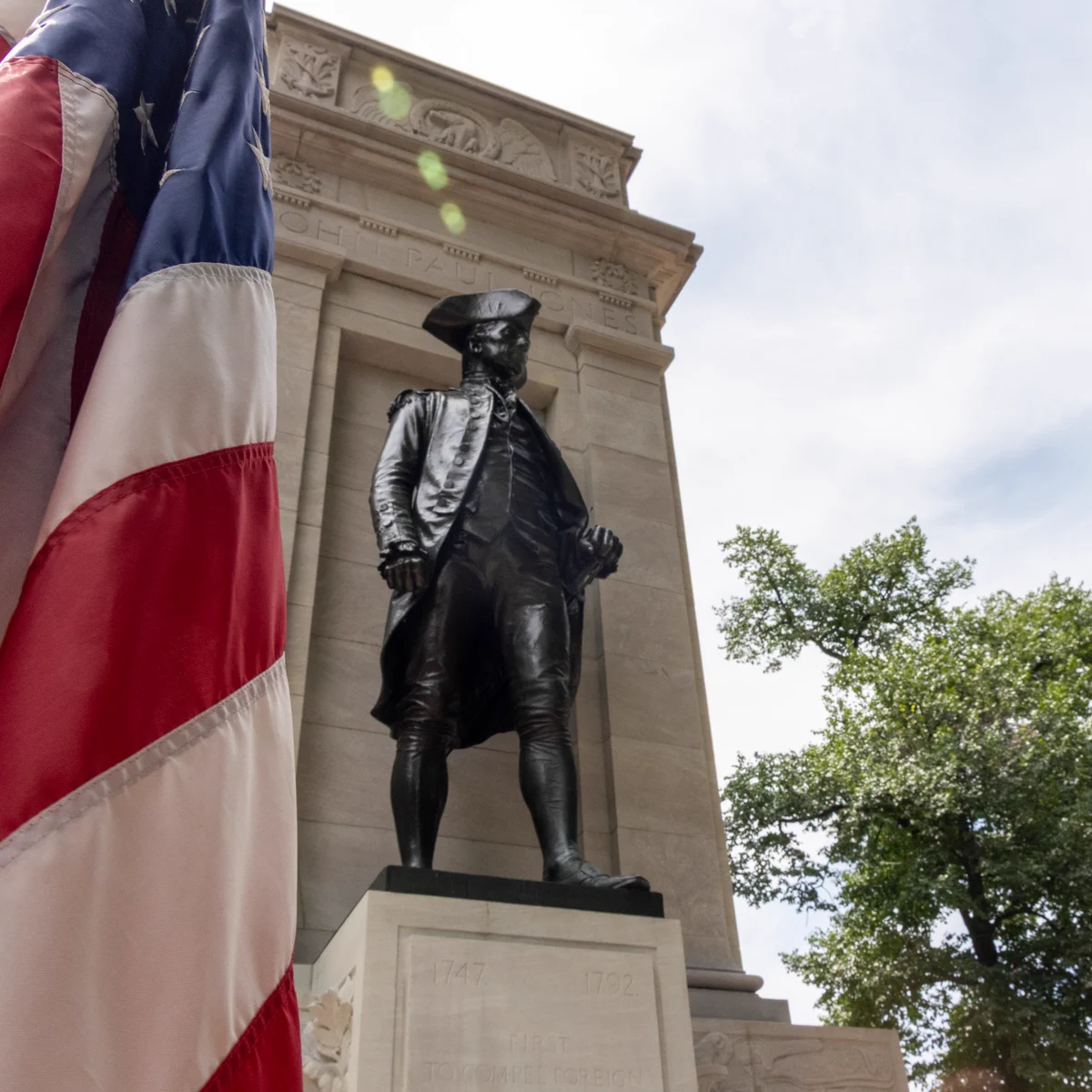 Bronze statue of naval commander John Paul Jones in period uniform and tricorn hat, standing before a tall stone pylon inscribed with his name and the dates 1747–1792, part of a memorial near the Tidal Basin in Washington, D.C.