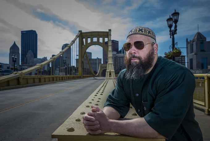 Sharif Bey stands on a bridge in Pittsburgh in the daytime with Pittsburgh's skyline visible in the background. He is wearing sunglasses and a patterned cap.