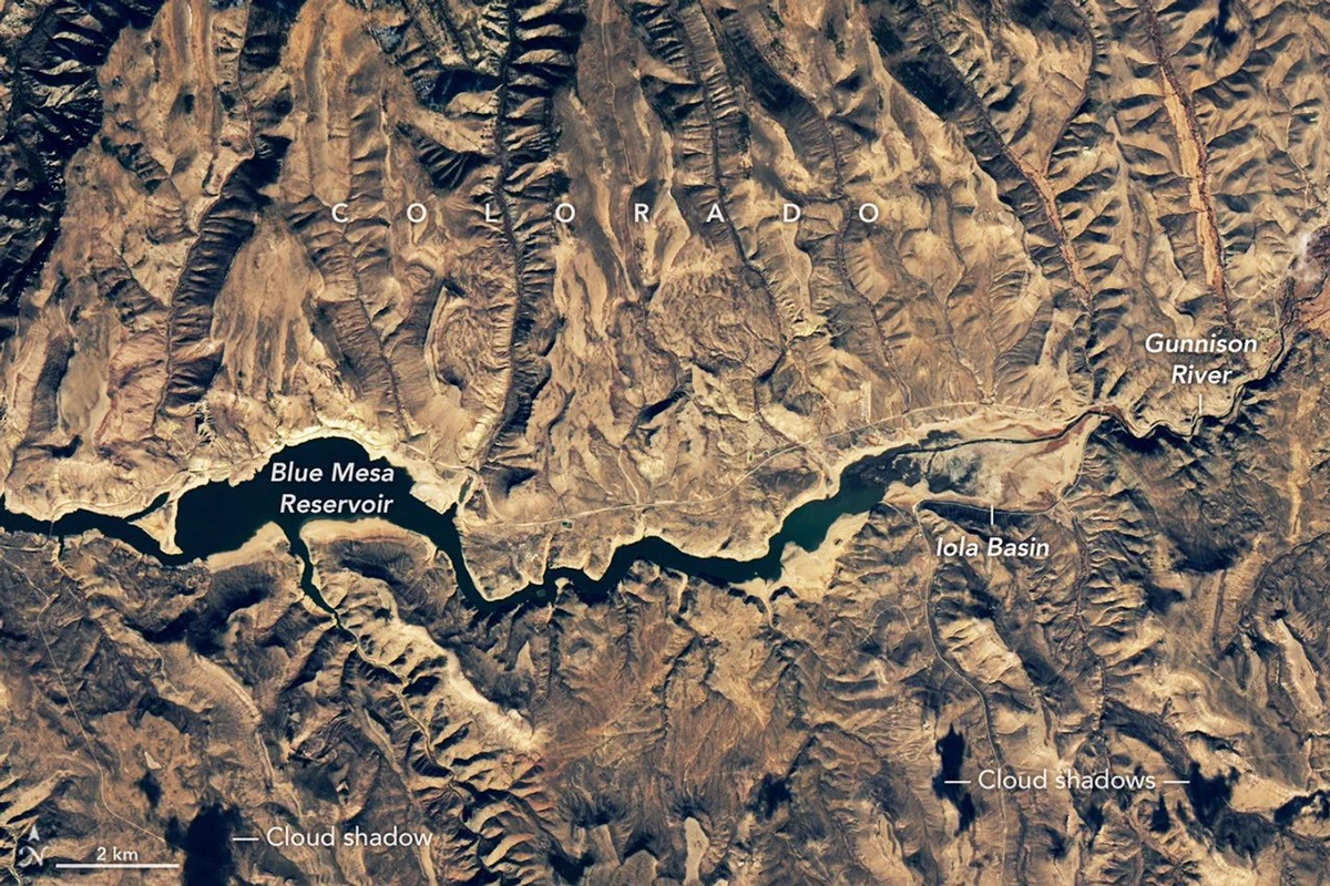 Landsat 8 satellite image showing the Blue Mesa Reservoir on November 17, 2021. The surrounding landscape is brown and mountainous. The reservoir, Iola Basin, and Gunnison River are dark blue and green, surrounded by paler land that was previously underwater when the water level was higher. Credit: NASA/USGS