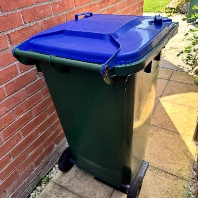 A green wheelie bin with a blue lid standing on a paved patio next to a red brick wall.