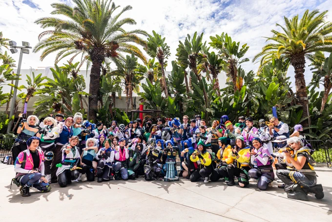 Large group of VALORANT cosplayers gathers for a group photo outdoors beneath palm trees during a community meetup at Anime Expo. 