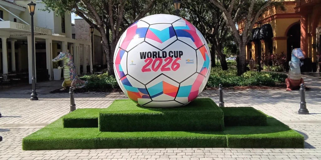 Large colorful soccer ball sculpture with 'World Cup 2026' text displayed outdoors on a green platform in a plaza.