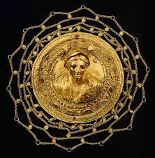Primer plano de una sección de una antigua redecilla para el cabello de origen griego helenístico, confeccionada con una intrincada malla de finas cadenas de oro entrelazadas de forma artesanal. La imagen muestra el delicado patrón de red geométrica y los eslabones dorados sobre un fondo oscuro, reflejando la avanzada orfebrería de la época.