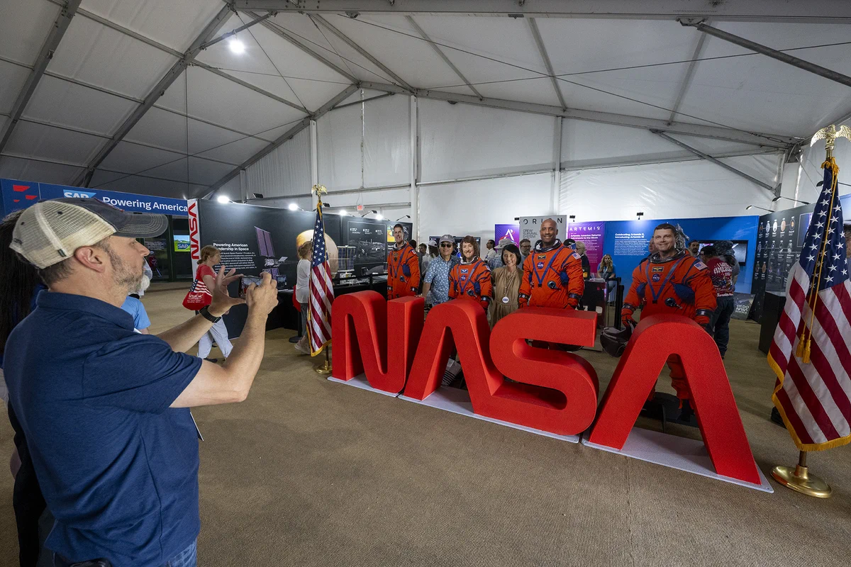 Visitors pose for a photo beside life-size astronaut cutouts wearing bright orange launch and entry suits inside NASA’s pavilion at the Great American State Fair. A visitor in the foreground takes the picture with a smartphone in front of large red NASA letters. American flags frame the display, while exhibits highlighting NASA technologies, space exploration, and the Artemis campaign line the pavilion walls.