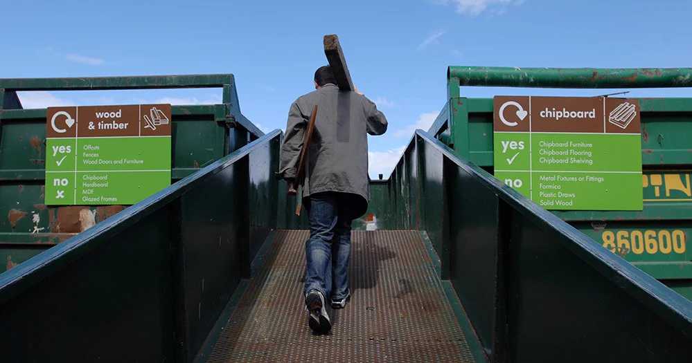 A man carrying timber walks up a ramp between two green recycling skips with signs for wood and chipboard disposal.