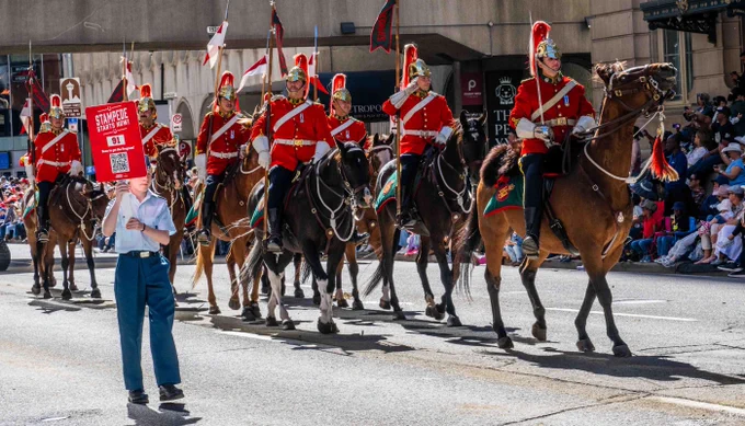 Mounted military soldiers in red uniforms and helmets riding horses during a city parade on a sunny day.