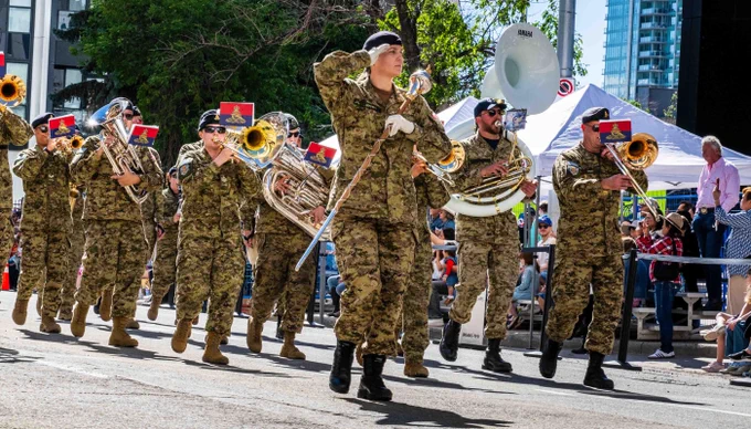 Group of soldiers in camouflage uniforms and black cowboy hats marching in formation during a parade with a seated crowd watching.