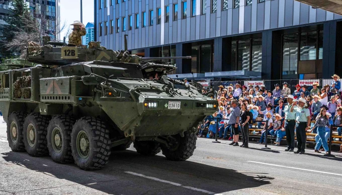 Military armored vehicle with soldiers driving past a crowd gathered on a city street during a public event.