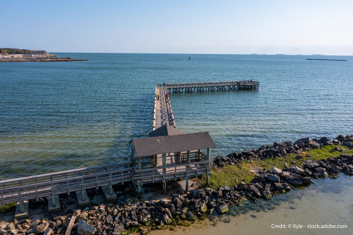 A rocky jetty crosses the frame in shallow Bay waters. A wooden walkway follows the jetty halfway, then turns and continues as an L-shaped wooden pier out into the water.