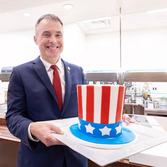 A person in a suit and red tie holds a cake shaped like a top hat with red and white stripes and white stars on a blue band. The background shows a kitchen setting.