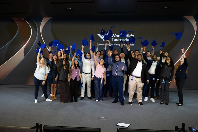 A diverse group of young adults in business casual attire celebrate, throwing blue graduation caps into the air. Behind them, a large screen displays \"Worldwide Technology Associate Academy Graduation\" and abstract patterns.