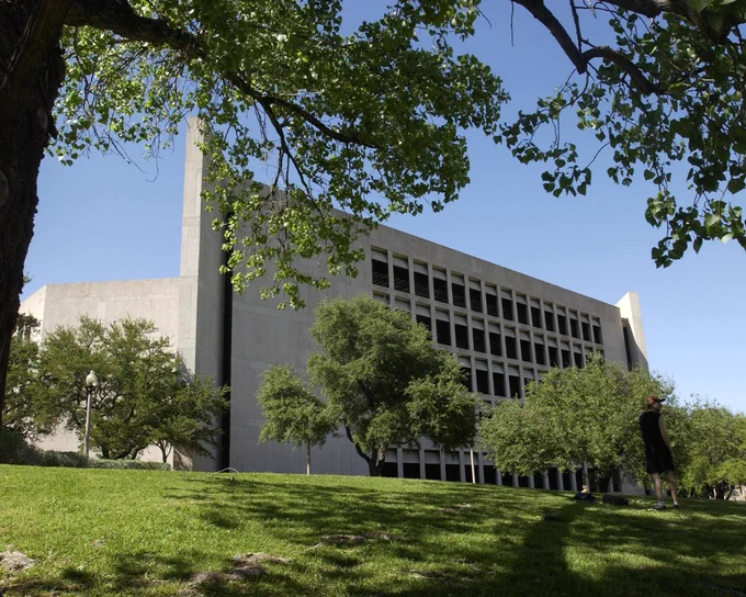 Modern concrete building with grid windows stands against a clear blue sky. A grassy hill with trees slopes up to it, dappled with shade. Overhead, green tree branches frame the scene. A person in a dark shirt & orange cap stands on the right.