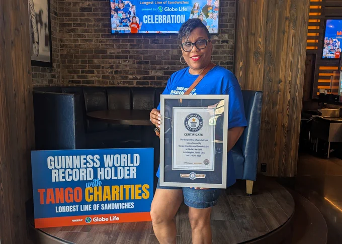 A person is standing indoors, holding a framed certificate in front of a brick wall. Next to them is a sign featuring the Guinness World Records logo and "Tango Charities," announcing a record for the longest line of sandwiches. The person is wearing glasses and a blue shirt.