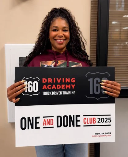 A person is smiling and holding a sign that reads, "160 Driving Academy Truck Driver Training - One and Done Club 2025.
