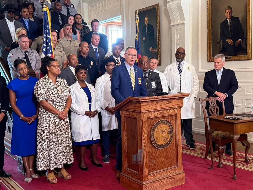 Secretary Jeffrey Smythe stands at a podium, speaking. A crowd of people stand behind him on the Executive Mansion staircase. Governor Josh Stein stands by a desk at the right side of the photo.