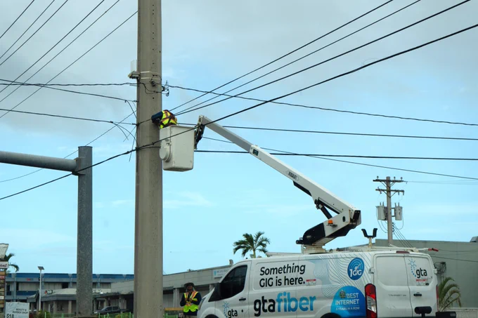 A utility worker in a safety vest works on a power pole from an elevated bucket truck. The truck is branded "gta fiber" and parked nearby, with another worker standing on the ground. Overhead power lines and a partly cloudy sky are visible in the background.