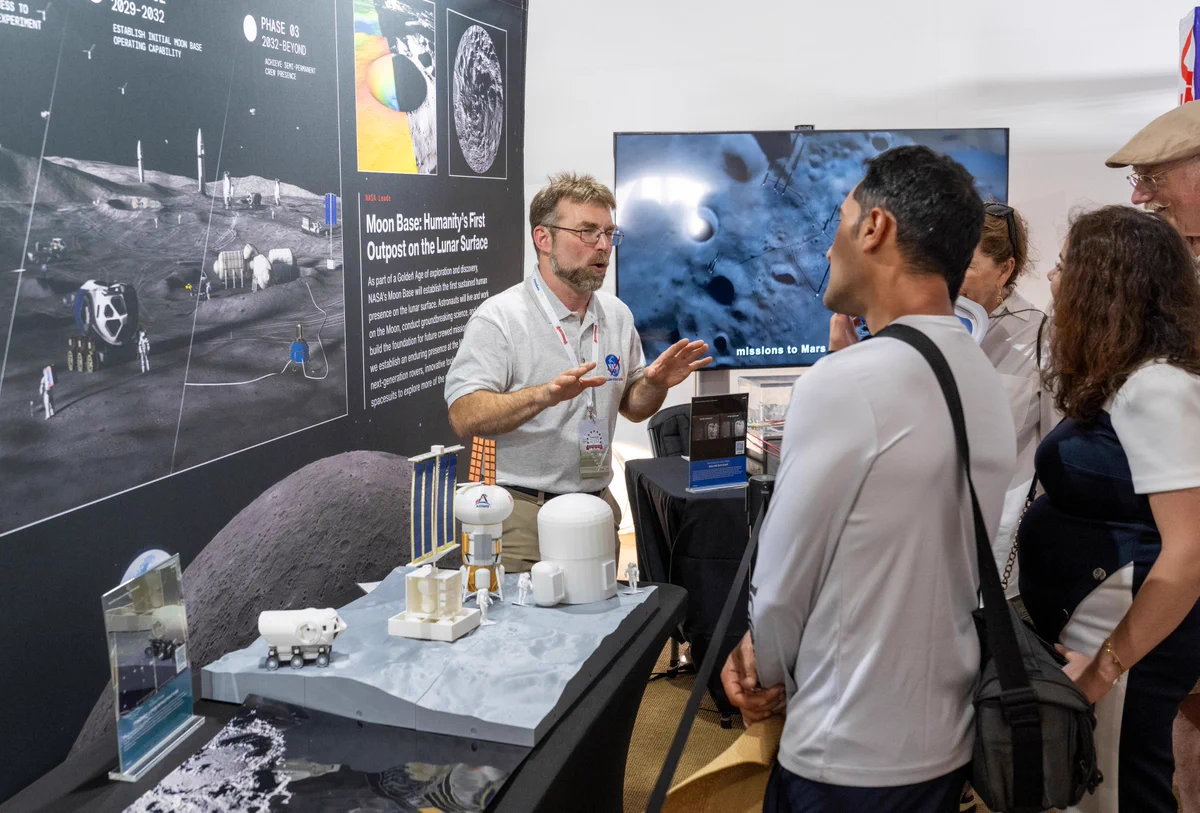 A NASA representative stands beside a tabletop PowerScape demonstration, explaining concepts to a small group of attendees. The PowerScape model features white scale models of lunar infrastructure—including habitats, a lander, and a rover—arranged on a gray terrain surface representing the Moon’s south polar region. Behind the display, a large graphic titled “Moon Base: Humanity’s First Outpost on the Lunar Surface” illustrates a future lunar settlement and exploration activities. Visitors listen as the presenter gestures toward the model during the discussion.