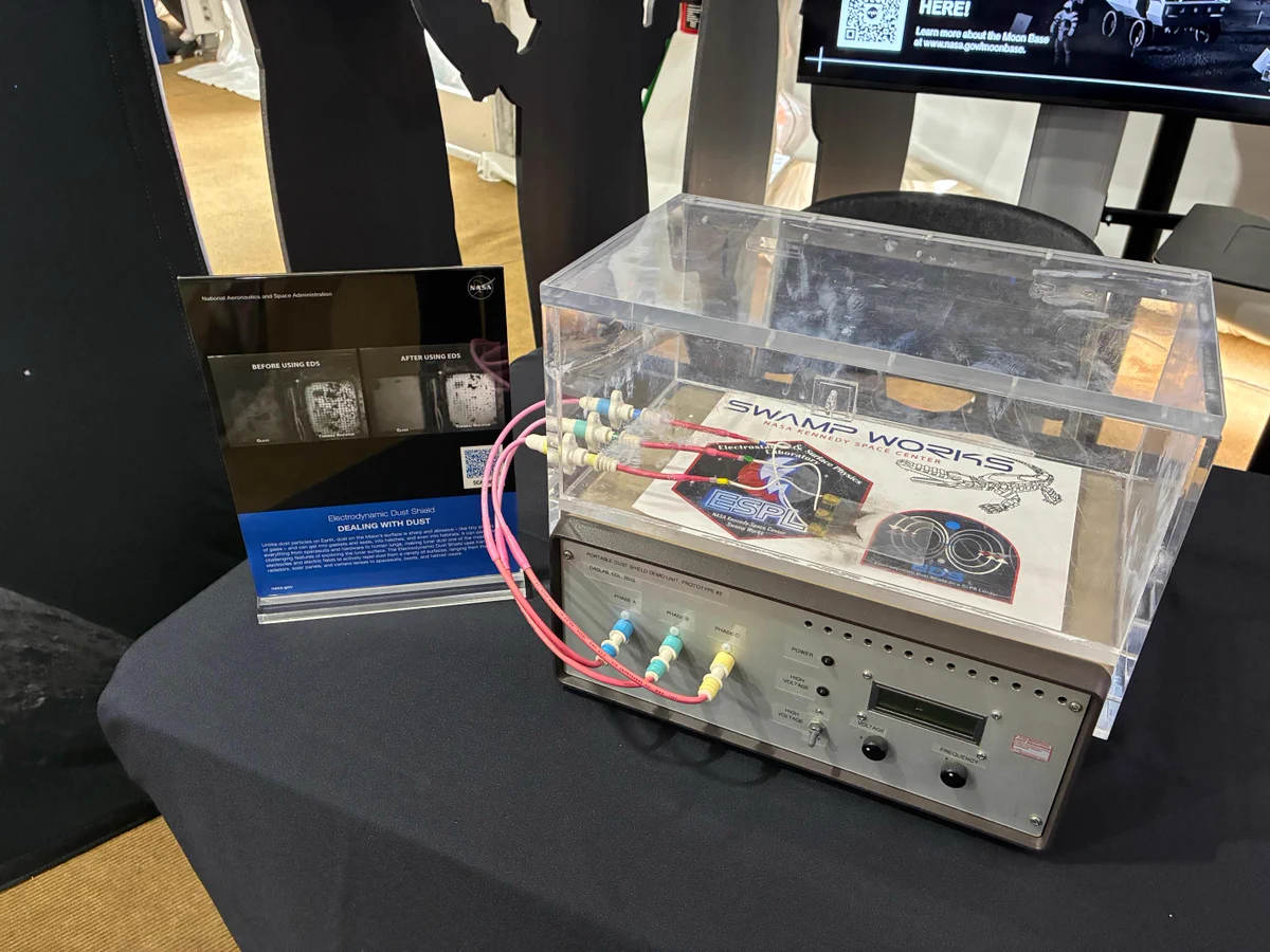A tabletop demonstration of NASA’s Electrodynamic Dust Shield technology is displayed at an exhibit. The prototype consists of a clear enclosure connected to a control unit with cables and power controls. An informational sign beside the display shows before-and-after images demonstrating how the technology removes lunar dust from surfaces. The dust mitigation system is designed to help protect equipment, habitats, spacesuits, and other hardware from abrasive Moon dust during future Artemis missions and long-term lunar exploration.