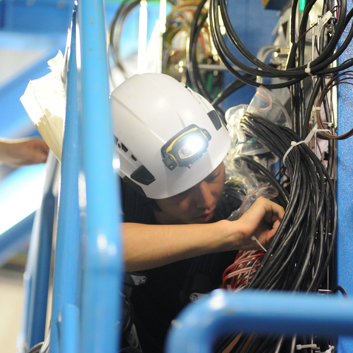 kek_ipns's tweet photo. 【HL-LHC：ATLASミューオントリガー③】
電子回路室と同時進行で、巨大な検出器本体からもケーブルを取り外します。最先端の実験現場ですが、ラックの裏側はまるで隙間のないジャングルです。

研究者たちは高所作業用のハーネスとヘッドライトを装備し、1本ずつケーブルを解きほぐしています。この確実で地道な手作業が、未来の新しい物理を支える強固な土台となります。（※動画は1.4倍速です）
