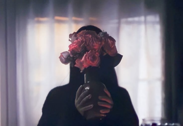 thevyrovisuals's tweet photo. While most people watch this scene in Obsession and assume "Wish Nikki" is just trying to look terrifyingly uncanny or use the roses as a twisted symbol of love, the real theory is much darker: the flowers are a biological shield. During her breakdown in the corner, "Real Nikki" was fighting violently to gain consciousness and scream for help, causing the One Wish Willow’s magic to literally glitch and fracture. She grabbed the vase because her face was actively melting and deforming into the blank, expressionless entity of the Willow itself, needing to hide the physical "rendering error" from Bear while the curse rebooted her human appearance. This is a direct nod to René Magritte’s painting The Son of Man (where an apple hides a face) because everything we see hides something else Wish Nikki is forcing Bear to look at "love" while hiding the grotesque reality underneath. Look closely at the puppet-like, jerky movements right before this and how her face goes completely blank and hollow the second she gets into bed; if you look between the stems, her eye is rolling into the back of her head. Curry Barker secretly hid a horrific body-horror transformation right in front of us, which begs the question: is this psychological horror, or did Bear’s subconscious definition of "love" literally create a faceless, mindless object?