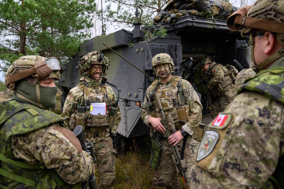 Four military members – two Canadians, two Norwegians – are in a circle, discussing. There’s a military vehicle behind them with a member in the back door and one on top.