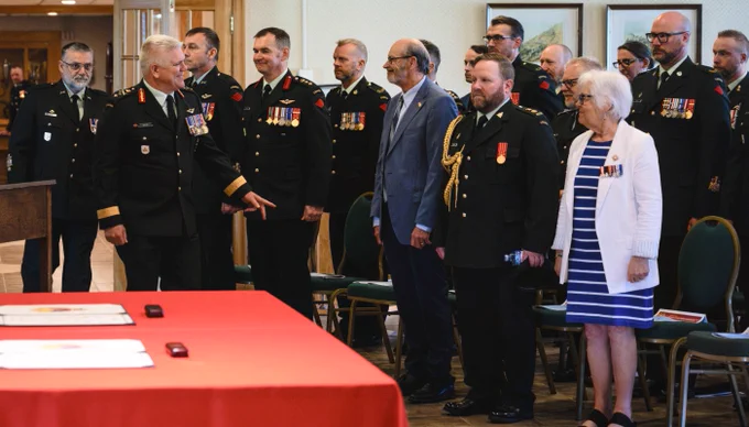 Group of military officers and civilians standing in a formal room, some wearing medals and uniforms, engaged in conversation.