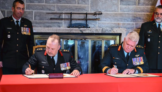 Two military officers in decorated uniforms sign documents at a red-covered table, flanked by two standing officers with Canadian flags behind them.
