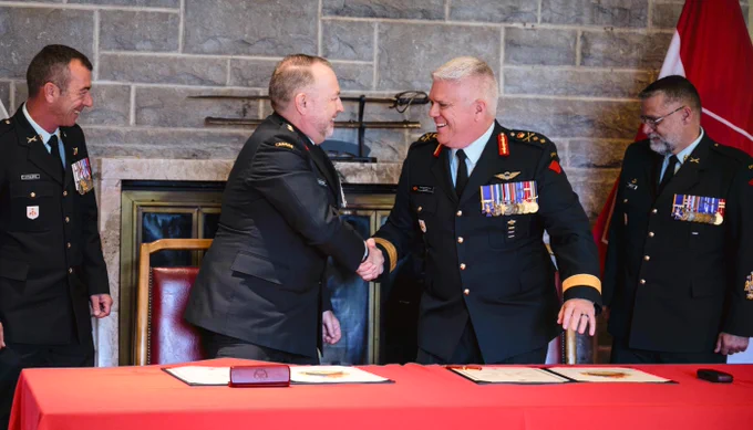 Two military officers in decorated uniforms shake hands over a red table with documents, while two other officers observe.