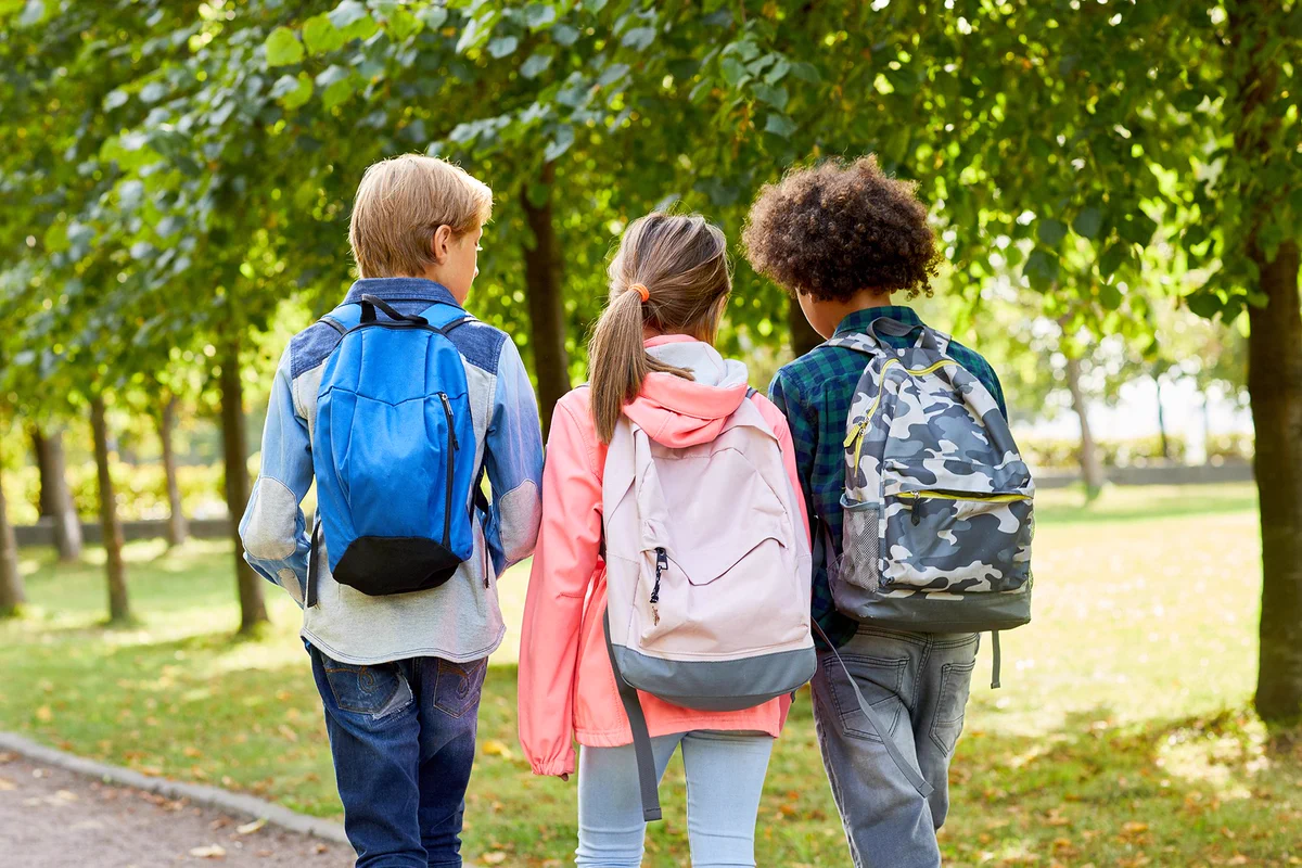 Three students wearing backpacks walk side by side along a path in a park. Green foliage fills the background.