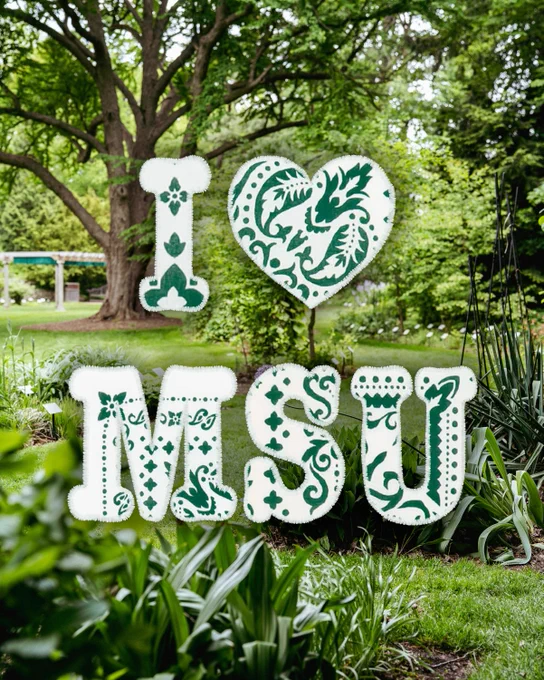 Large decorative white letters and a heart, filled with intricate green patterns, spell \"I \u2764\uFE0F MSU\". They are placed in a vibrant, lush green garden with a large tree in the background and various bushes and plants surrounding them.