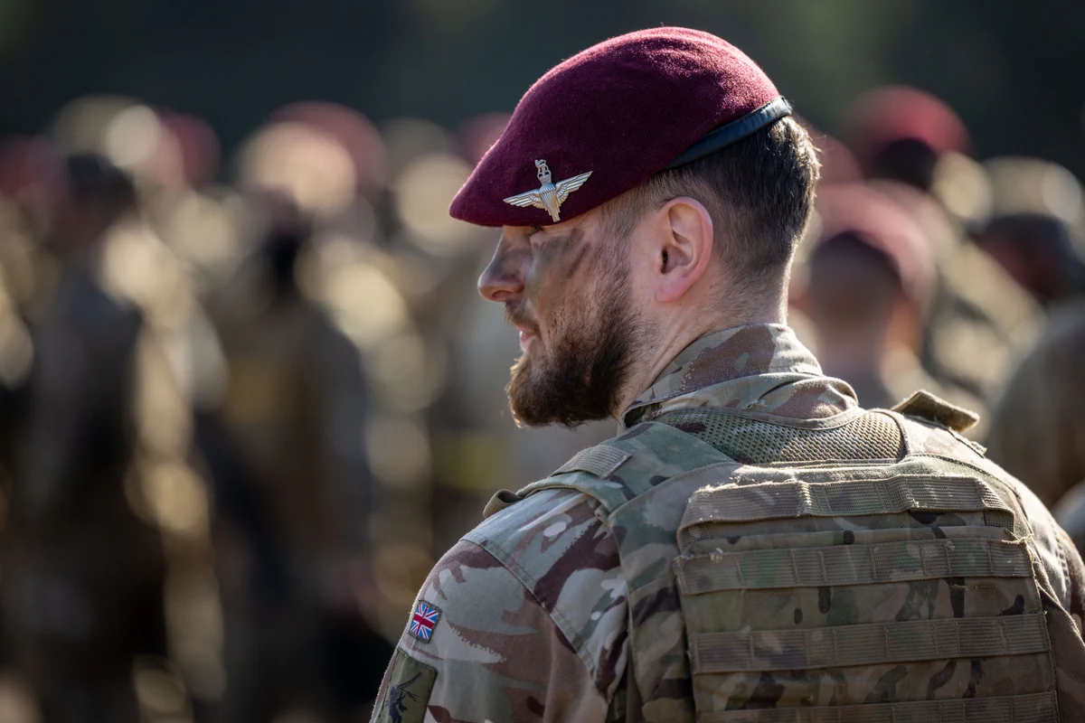 A person in military camouflage uniform and a maroon beret with a winged badge stands among a group of similarly dressed individuals. The uniform bears a patch of the British flag on the shoulder.