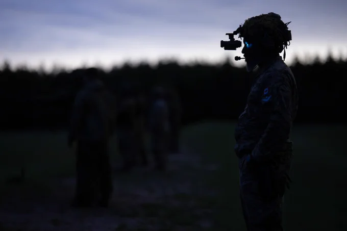 Silhouette of a soldier wearing night vision goggles with blurred figures in the background.