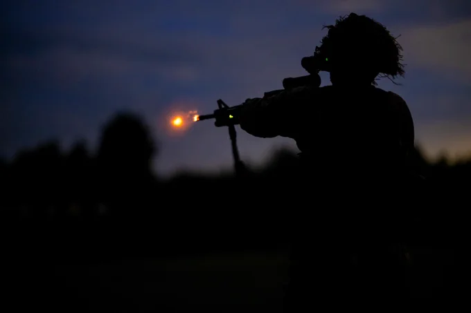Silhouette of a soldier firing a rifle at dusk with a visible muzzle flash.
