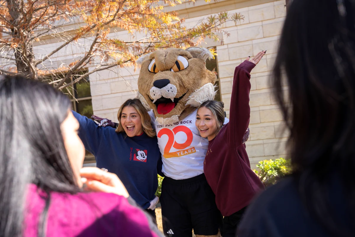 Two people pose excitedly with a large bobcat mascot wearing a shirt celebrating 20 years outside a building on a sunny day.