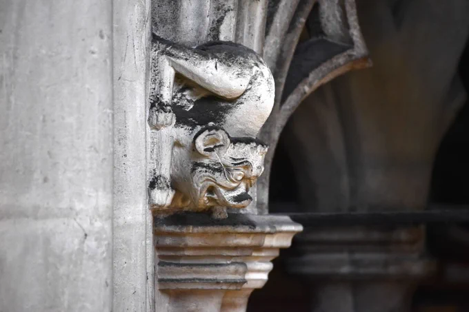 A stone sculpture with a grotesque, crouching figure. Its face features exaggerated, curled expressions, and it clings to a decorative architectural structure. The surface shows signs of age, with weathered textures and darkened areas.