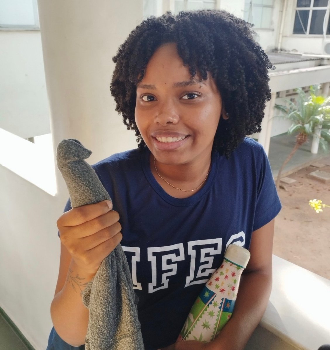 acervoestersoar's tweet photo. Ester Soares usando camisa azul escrito IFES segurando um pano em formato de pênis e sua garrafa térmica colorida da farm sorrindo como se tivesse sido promovida a funcionária do mês transbordando carisma https://t.co/MUJkZunPyQ