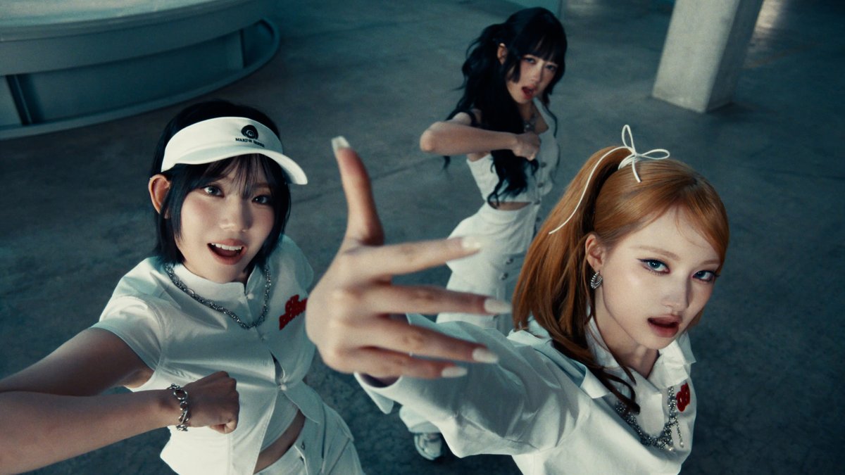 Three women in white outfits pose dynamically with pointing gestures in what appears to be a K-pop music video still, with one wearing a white visor.