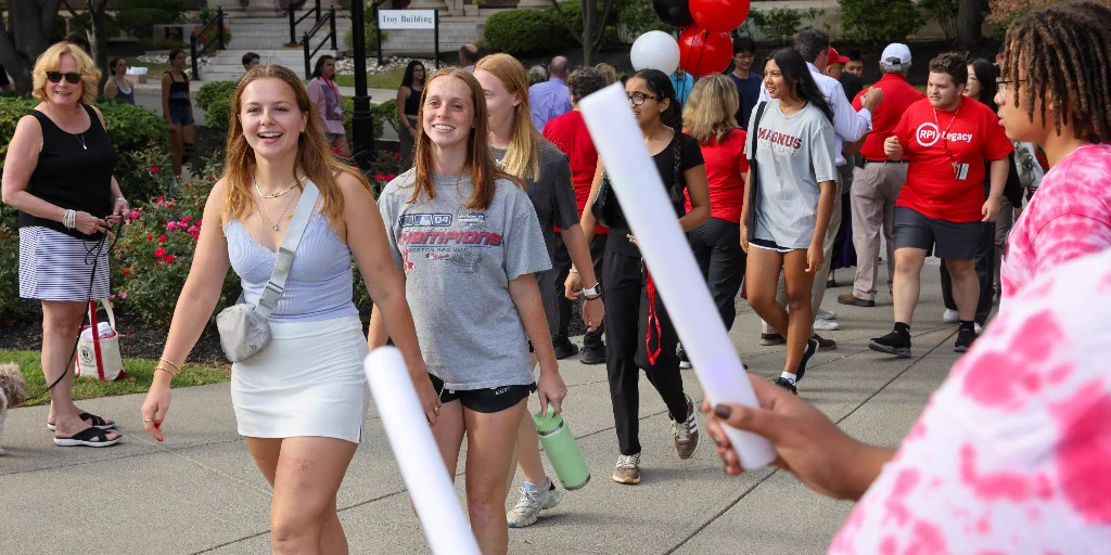 Students walking in a group on the Rensselaer Polytechnic Institute campus, with red and black balloons in the background. They are casually dressed, and a building with columns is visible in the background.