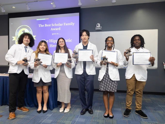 Six people standing in a room, all wearing white coats and holding certificates and small trophy figures. A screen in the background displays a presentation slide that reads "The Best Scholar Family Award goes to..."