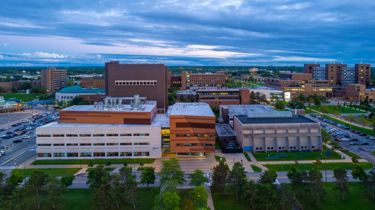 Aerial view of a large campus under a cloudy, evening sky. Numerous academic & modern buildings of brick, stone, & patterned facades line green spaces, roads, & parking lots filled with cars. 'Barbara and Herb Davis Hall' is visible on one. Lights glow from some windows.