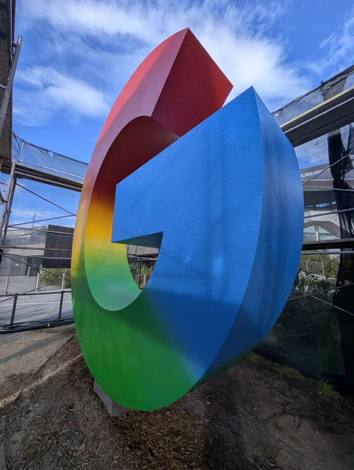 A low-angle, close-up shot of the giant, freshly painted multicolor Google "G" statue against a blue sky.
