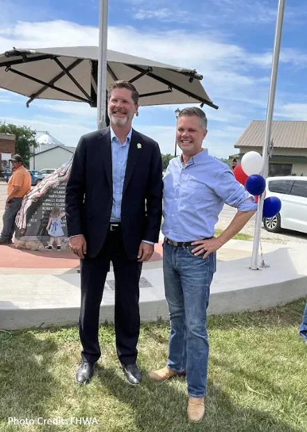 Admin. McMaster and Rep Nunn stand outdoors on grass in front of a podium with balloons and a large umbrella. One wears a suit, and the other wears a light shirt and jeans. A building is visible in the background. Photo credit: FHWA.

