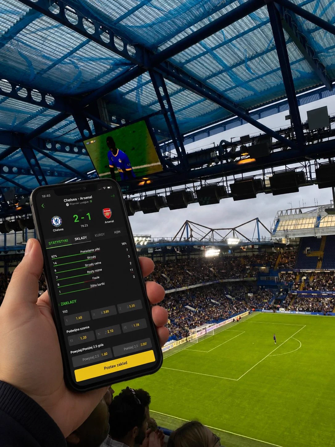 A football fan inside a packed Premier League stadium, holding a smartphone showing a live betting interface with hundreds of active markets. Crowd roaring in the background, floodlights on, green pitch visible below. The atmosphere is electric — this is matchday, and every second counts.