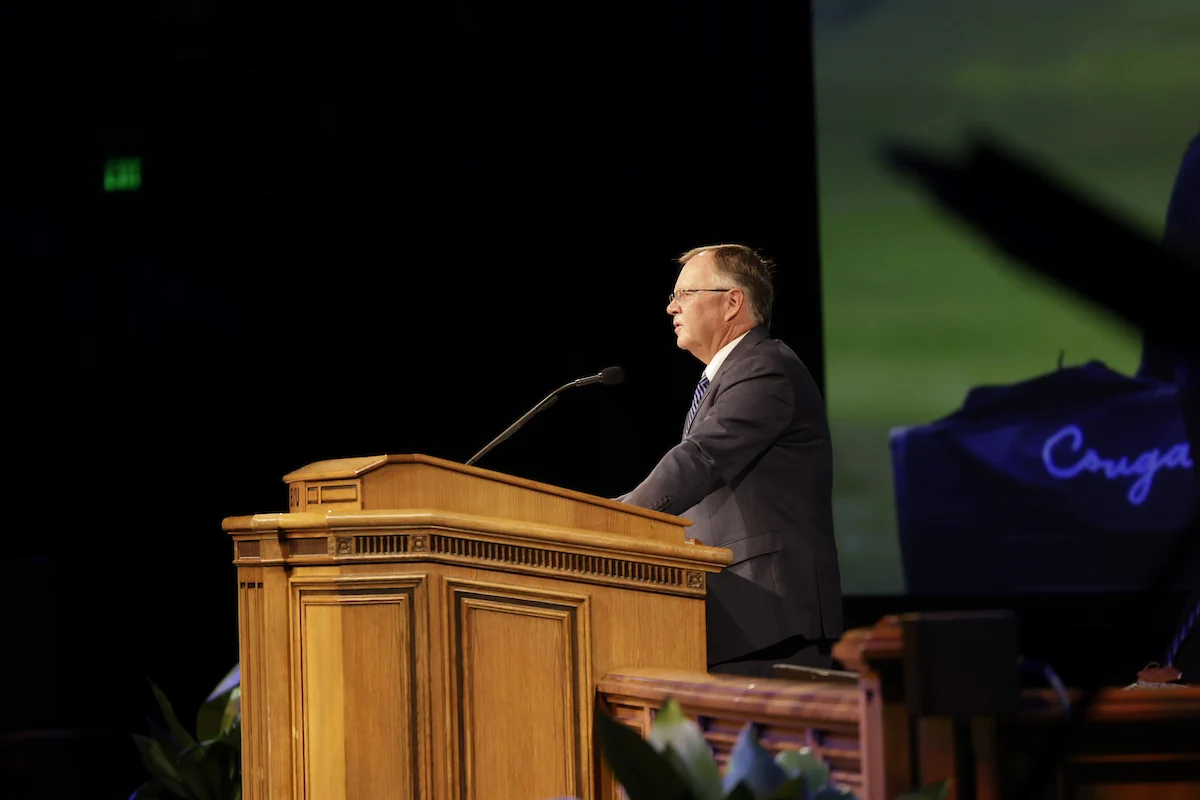 A side profile of Coach Bruce Brockbank speaking at a podium for BYU Devotional, July 2026