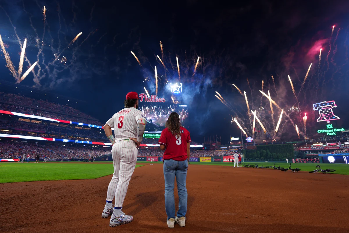 Bryce Harper stands with a fan on the field as fireworks go off in the distance. He wears a home white Phillies uniform as the fan wears a Harper T-shirt jersey.