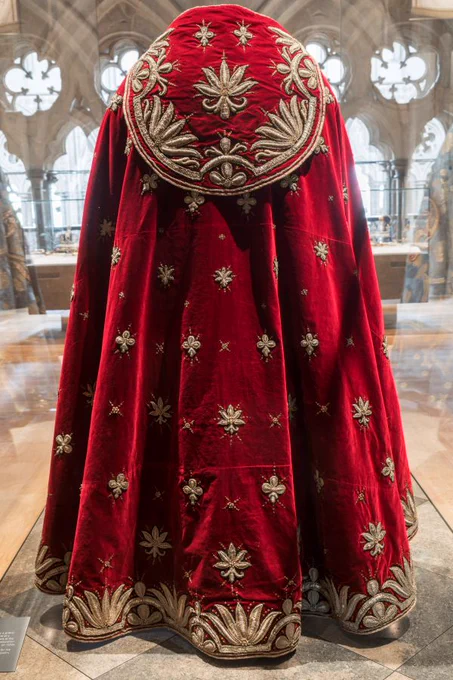 A richly embroidered red velvet cope is displayed, featuring intricate gold floral and leaf motifs. The cope has a circular shoulder cape with elaborate decoration and a full-length drape that pools elegantly on the floor. The backdrop reveals ornate arched windows, suggesting an exhibition setting.