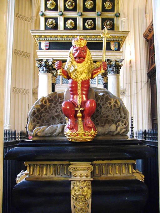 A vibrant red and gold heraldic lion statuette sits atop a decorative pedestal, holding a scepter. The lion features a detailed mane and crown, and is placed against an ornate background with intricate stone and metallic details in a grand architectural setting.