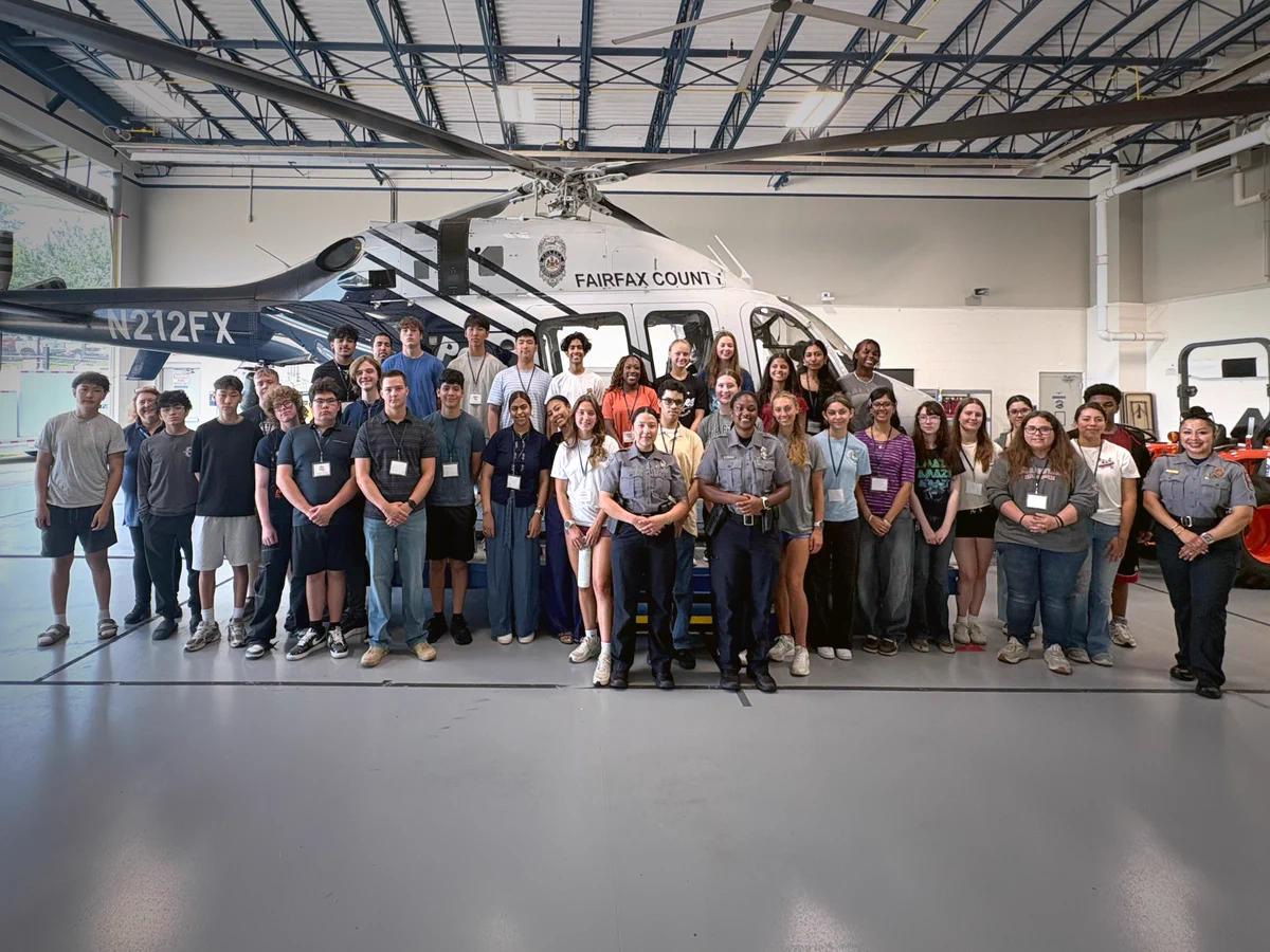 Group of diverse young people and officers posing in front of a Fairfax County police helicopter inside a hangar.