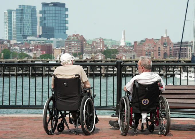 Two individuals in wheelchairs are sitting on a brick promenade by the water, looking towards a cityscape. There are buildings and a marina in the background.